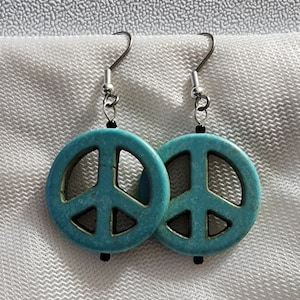 Puede incluir: Un par de pendientes de signo de paz turquesa con ganchos plateados. Los signos de paz están hechos de un material texturizado y tienen una cuenta negra en la parte inferior de cada pendiente.