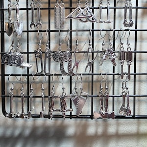 May include: A collection of silver-colored earrings in various tool shapes, including shovels, wrenches, and saws, displayed on a black wire grid. The earrings are detailed and hang from silver hooks, showcasing a unique, industrial theme.