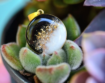 May include: A round pendant with a black and white resin design. The pendant is set in gold and features gold flakes and white specks.