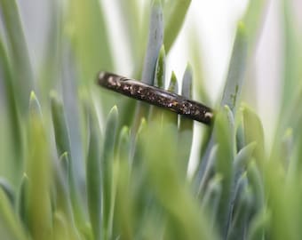 May include: A dark ring with a speckled design rests among green succulent leaves. The ring appears to be made of a dark metal with a textured surface. The background is blurred, highlighting the ring.