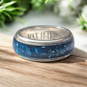 May include: A silver-toned ring featuring a wide band with a dark blue, textured inlay of crushed material. The interior of the metal band is engraved with the text MAX 11-11-2022, showing a smooth, polished finish on the edges of the ring.