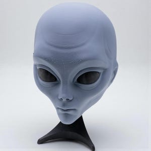 Op de afbeelding: Een lichtblauw alienmasker met grote zwarte ogen en een glad, afgerond hoofd. Het masker is gemonteerd op een zwarte standaard, die de gedetailleerde gelaatstrekken benadrukt. Een uniek item voor kostuum of display.