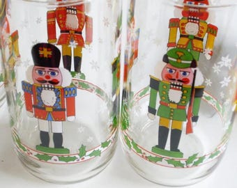 May include: Clear glass drinking glasses with colorful nutcracker soldier designs. The glasses feature red, green, and yellow nutcrackers with intricate details, set against a backdrop of snowflakes and a holly border.