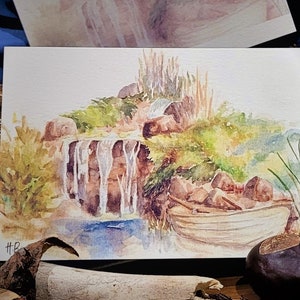 Könnte beinhalten: Aquarellmalerei eines kleinen Wasserfalls, der über Felsen und Grün herunterstürzt, mit einem Holzboot, das auf dem Wasser darunter liegt. Das Gemälde hat eine leichte, luftige Atmosphäre und ist in einem weißen Passepartout gerahmt.