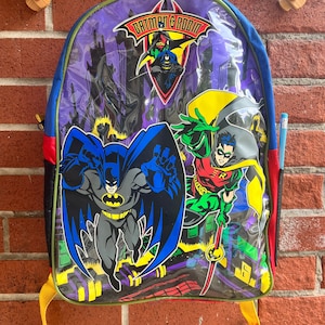 Puede incluir: Una mochila de Batman y Robin de color morado, azul, rojo y amarillo con un panel frontal de plástico transparente. El panel frontal presenta un gráfico de Batman y Robin en un paisaje urbano. La mochila tiene un tirador de cremallera amarillo y una correa amarilla.