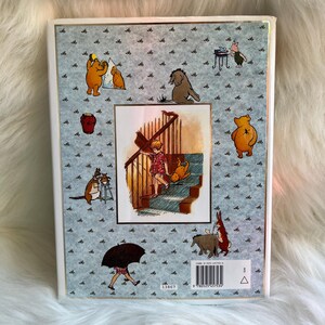 Puede incluir: Un libro de tapa dura con ilustraciones de Winnie the Pooh y sus amigos. La portada tiene un fondo azul claro con peque&ntilde;os gr&aacute;ficos de abejas negras. La imagen central muestra a un ni&ntilde;o y un perro en unas escaleras. El ISBN del libro es visible.