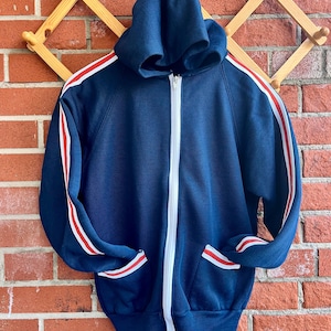 Puede incluir: Una sudadera con capucha azul marino con rayas blancas y rojas en las mangas y los bolsillos.