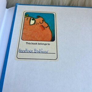 Puede incluir: Un libro abierto con lomo azul y p&aacute;ginas blancas. Una pegatina en la p&aacute;gina dice "Este libro pertenece a Heather Balfour" encima de una ilustraci&oacute;n de dibujos animados de una criatura redonda y naranja con fondo azul.
