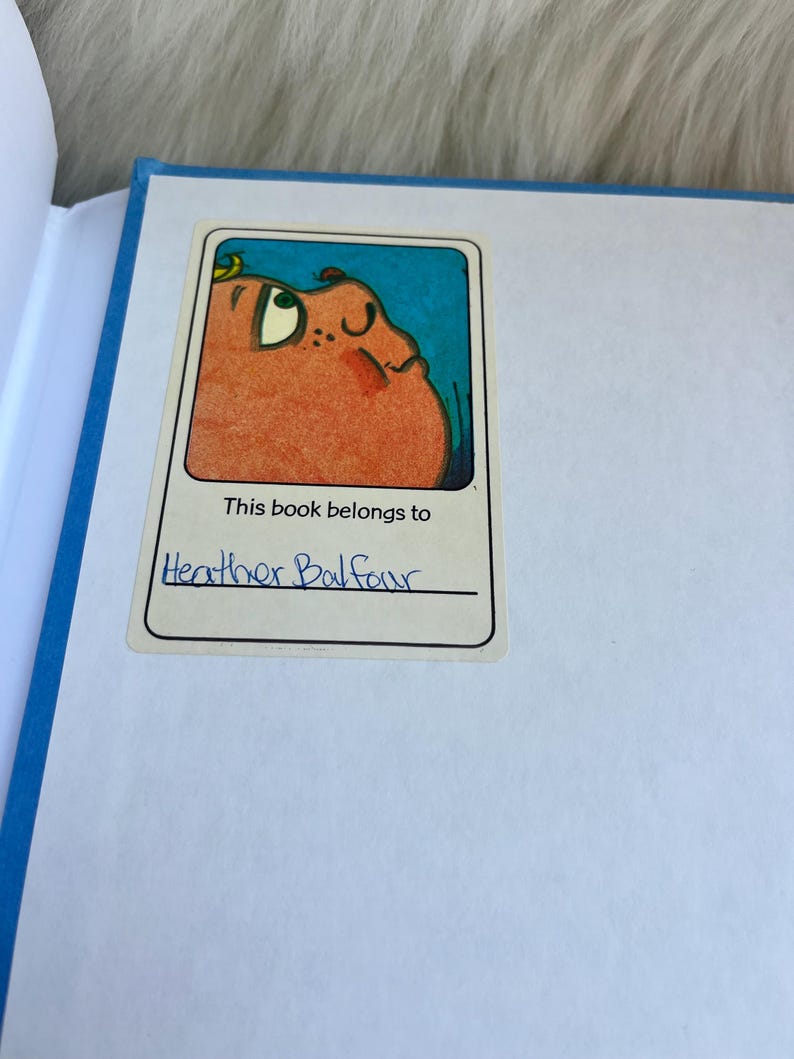 Puede incluir: Un libro abierto con lomo azul y p&aacute;ginas blancas. Una pegatina en la p&aacute;gina dice "Este libro pertenece a Heather Balfour" encima de una ilustraci&oacute;n de dibujos animados de una criatura redonda y naranja con fondo azul.