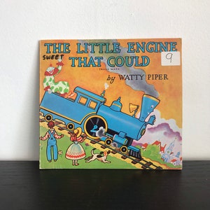 Könnte beinhalten: Ein Vintage-Kinderbuchcover mit einer blauen Lokomotive, die einen Waggon einen Hügel hinaufzieht. Der Buchtitel lautet "The Little Engine That Could" mit den Worten "Sweet" und "Trade Mark" über dem Titel. Das Buch ist Nummer 9 in einer Reihe und wurde von Watty Piper geschrieben.