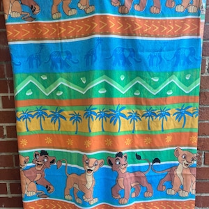 Puede incluir: Una colorida toalla de playa con el tema de El Rey León. La toalla presenta leones de dibujos animados, elefantes, jirafas y palmeras. El diseño incluye rayas naranjas, azules, verdes y amarillas con patrones de flechas.