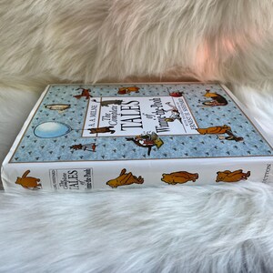 Puede incluir: Un libro de tapa dura titulado "The Complete Tales of Winnie-the-Pooh" de A.A. Milne. El libro tiene una cubierta azul claro con ilustraciones de Winnie the Pooh y sus amigos. El libro est&aacute; sobre una superficie blanca y esponjosa.