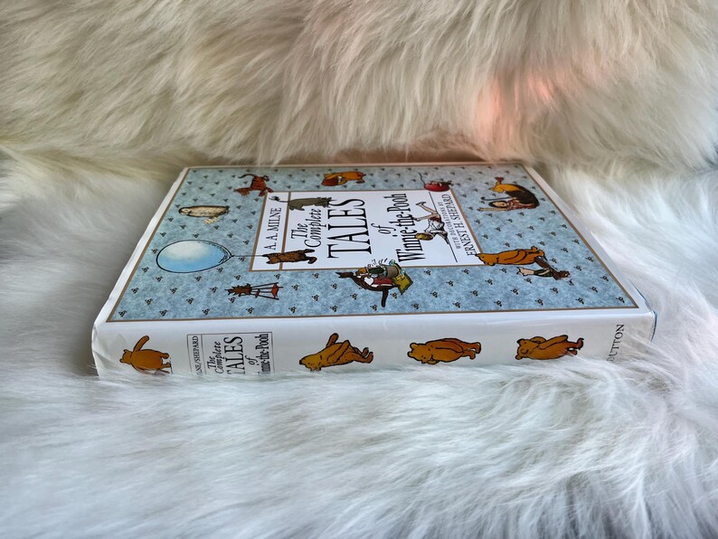 Puede incluir: Un libro de tapa dura titulado "The Complete Tales of Winnie-the-Pooh" de A.A. Milne. El libro tiene una cubierta azul claro con ilustraciones de Winnie the Pooh y sus amigos. El libro est&aacute; sobre una superficie blanca y esponjosa.