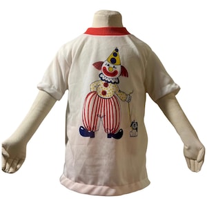 Puede incluir: Camiseta blanca con cuello y mangas rojas, con un gráfico de payaso colorido. El payaso tiene pantalones a rayas rojas y blancas, zapatos azules y un sombrero amarillo. El payaso sostiene una correa atada a un perro pequeño.