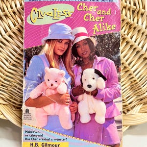Op de afbeelding: Een paperback boek met de titel "Clueless Cher and Cher Alike" met een roze en paarse omslag. De omslag toont twee vrouwen die knuffels vasthouden. De tekst "Based on the ABC-TV Prime Time Series" staat bovenaan.