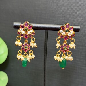 May include: Gold-toned floral earrings with ruby-colored petals, white accents, and dangling pearls. Each earring features a green teardrop-shaped stone at the bottom. The earrings are displayed on a black stand against a dark background.