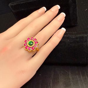May include: A gold-toned ring featuring a flower design. The ring has a central green stone surrounded by pink petals. The ring is displayed on a hand with manicured nails.