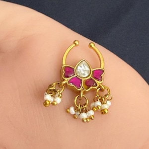 May include: A gold-toned nose ring featuring a central teardrop-shaped clear stone surrounded by four magenta-colored petal accents. The piece includes three dangling charms, each composed of small white beads and tiny gold-toned spheres.