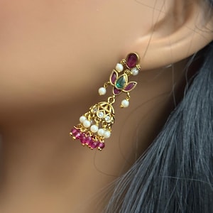 May include: Gold-toned dangle earrings with a floral design. The earrings feature ruby-colored and emerald-colored gemstones, small white pearls, and a bell-shaped cluster of ruby-colored stones. The earrings are worn on a person's ear.