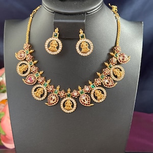May include: A gold-toned necklace and earring set displayed on a black stand. The necklace features a series of circular and paisley-shaped pendants adorned with red, green, and gold accents. The earrings mirror the circular design. The jewelry is likely for special occasions.