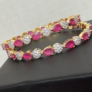 May include: Two gold-toned bangles with alternating pink teardrop-shaped and round, clear, sparkling stones. The bangles are displayed on a dark surface, showcasing the jewelry's design and color contrast.