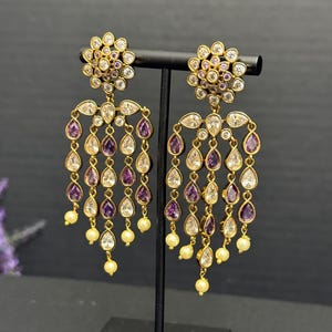 May include: Gold-toned chandelier earrings with a floral design at the top. The earrings feature clear and purple teardrop-shaped stones, with small pearl-like beads at the bottom. The earrings are displayed on a black stand.
