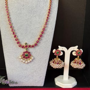 May include: A gold-toned necklace and earring set. The necklace features a series of red gemstones and a pendant with a floral design, green accents, and white pearls. The earrings have a similar design, with red gemstones and white pearls.