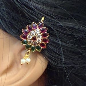 May include: A gold-toned earring with a floral design, featuring red, purple, green, and clear gemstones. Two small white pearls dangle from the bottom. The earring is worn on an ear against a backdrop of dark hair.