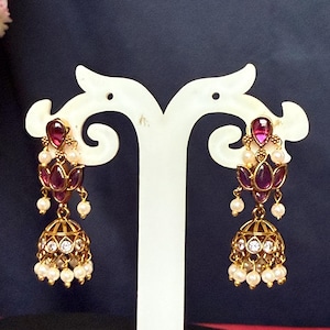 May include: Gold-toned earrings with a floral design, featuring ruby-colored gemstones and small white pearls. The earrings have a bell-shaped bottom with clear crystals. They are displayed on a white stand against a dark background.