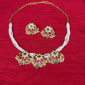 May include: A gold-toned necklace and matching earrings set. The necklace features three floral pendants with green and pink gemstones, strung on a white beaded strand. The earrings have a similar floral design. The jewelry set is displayed on a red background.