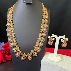 May include: A gold necklace with red and green stones, and matching earrings. The necklace is on a black display stand and the earrings are on a white display stand.