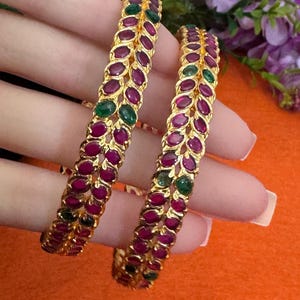 May include: Two gold bangles with alternating ruby red and emerald green gemstones. The bangles are held up against a bright orange background. The gemstones are oval-shaped and set in a gold band.