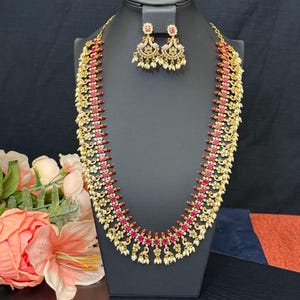 May include: A gold-toned necklace and matching earrings set. The necklace features alternating red and white stones, with small white pearl-like accents. The earrings have a similar design. The jewelry is displayed on a black mannequin against a dark background.