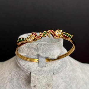 May include: A gold-colored bangle bracelet with a decorative top featuring red, green, and white gemstones. The bracelet is displayed on a gray, plush surface against a dark background. The gemstones are set in a detailed, ornate design.