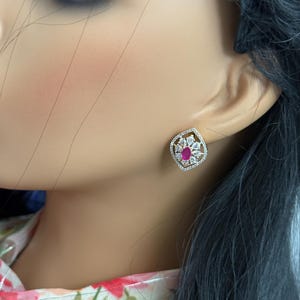 May include: A stud earring featuring a central oval-shaped red stone surrounded by a cluster of small, clear, pear-shaped stones. The entire arrangement is set within a gold-toned, diamond-shaped frame lined with tiny, clear, sparkling stones.