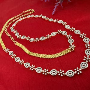 May include: A gold-toned necklace with a delicate design, featuring a series of floral and teardrop-shaped elements encrusted with small, sparkling white stones. The necklace is displayed on a red surface.