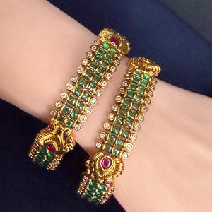 May include: Two gold bangles with green and clear gemstones and red accents. The bangles have an ornate design with a repeating pattern of green square stones and clear round stones. The ends of the bangles have a detailed, decorative design.