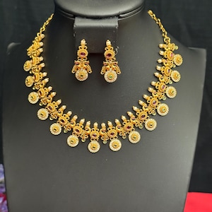 May include: A gold-toned necklace and earring set displayed on a black mannequin. The jewelry features intricate floral and circular designs with red and white accents. The necklace and earrings are likely made of metal.
