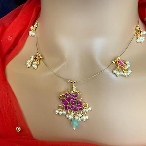 May include: A gold necklace with a pink and red pendant featuring a design of a deity. The pendant is adorned with white pearls and a light blue stone. The necklace also has two smaller pendants with white pearls.