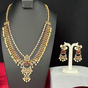 May include: A gold-toned necklace and earring set displayed on a black mannequin. The necklace features multiple layers with red and white accents, and a pendant with a red stone. The earrings have a similar design.