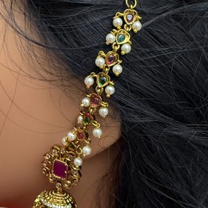 May include: Gold-toned earring with a floral design, featuring a central square ruby-colored stone and a dangling bell-shaped element. The earring is adorned with small pearls and colorful gemstones, creating an elegant and ornate appearance.