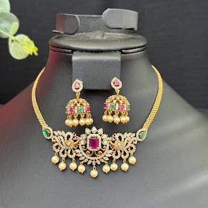 May include: A gold-toned necklace and earring set. The necklace features a central square ruby-colored stone, surrounded by clear stones and dangling pearls. The earrings have a similar design with ruby-colored stones, green and red accents, and dangling pearls.
