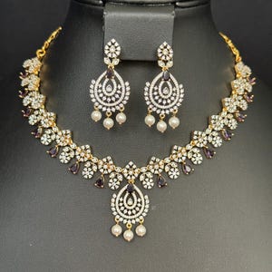 May include: A gold-toned necklace and earring set. The necklace features a floral design with white and purple stones, and a teardrop pendant. The earrings are teardrop-shaped with white stones and dangling pearls. The jewelry is displayed on a black stand.