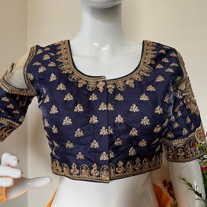 May include: A navy blue embroidered blouse with gold thread detailing. The blouse has a scoop neckline and short sleeves with cutouts at the shoulders.