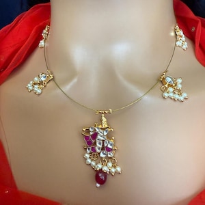 May include: A gold necklace with a pink and white pendant featuring a geometric design. The pendant is adorned with small pearls and a single red gemstone. The necklace has two smaller pearl and gemstone accents on either side of the pendant.