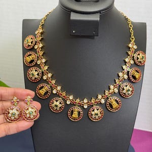 May include: Gold-toned necklace and earring set. The necklace features circular pendants with red, green, and white gemstones, and gold figures. The earrings mirror the necklace design. The jewelry is displayed on a black mannequin.