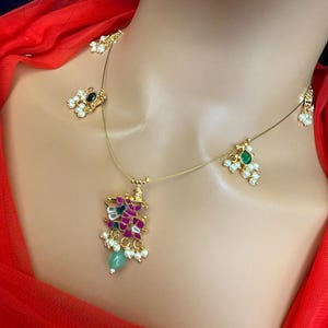 May include: A gold necklace with a pendant featuring a pink and green floral design. The pendant is surrounded by small white pearls and green stones. The necklace also has two smaller pendants with green stones and white pearls.