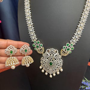 May include: A dazzling jewelry set featuring a necklace and matching earrings. The necklace has a floral design with clear and green stones, and a large pendant with dangling pearls. The earrings have a similar design with a pearl drop.