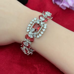 May include: A silver-toned bracelet with a central rectangular red gemstone, surrounded by smaller white and red stones. The bracelet features a symmetrical design with alternating red and white accents, creating a sparkling, elegant look.