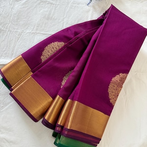 May include: A purple silk saree with a gold border and intricate gold designs. The saree is folded and displayed on a white surface.